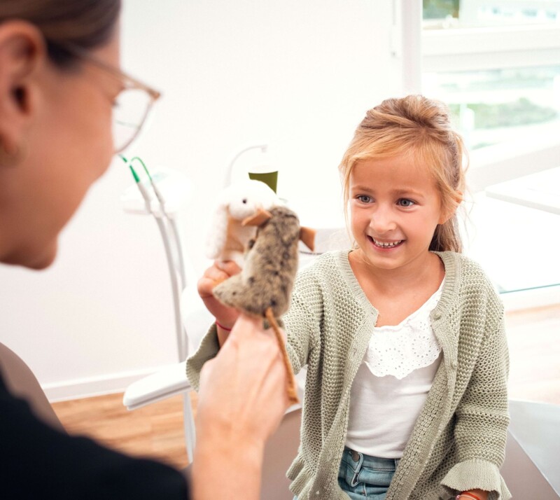 Ein kleines Mädchen sitzt mit Frau Riekert im Behandlungszimmer, mit Fokus auf dem Mädchen. Die beiden spielen zusammen mit Fingerpuppen in Form von kleinen Mäusen.