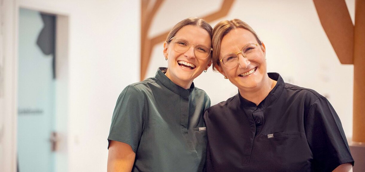 Kinderzahnärztin Elisa Riekert (links) und Ursula Schönleber (rechts) stehen Kopf an Kopf im Empfang Ihrer Praxis Zahnwald in Mühlacker und grinsen sympathisch.