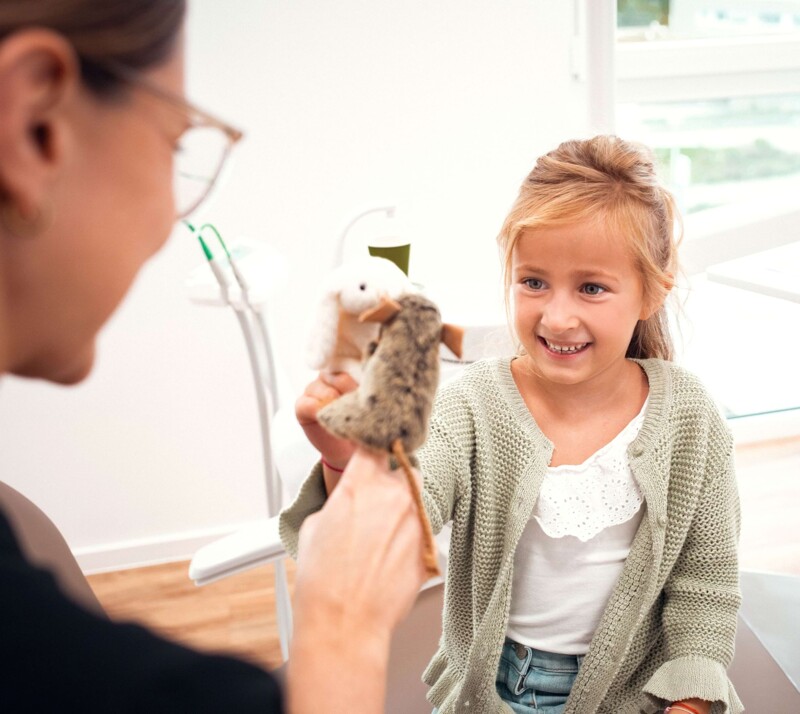Ein kleines Mädchen sitzt mit Frau Riekert im Behandlungszimmer, mit Fokus auf dem Mädchen. Die beiden spielen zusammen mit Fingerpuppen in Form von kleinen Mäusen.