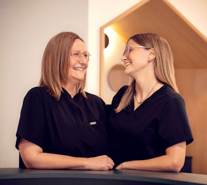 Frau Riekert (rechts) und Frau Schönleber (links) stehen lächelnd an der Rezeption ihrer Kinderzahnarztpraxis Zahnwald in Mühlacker