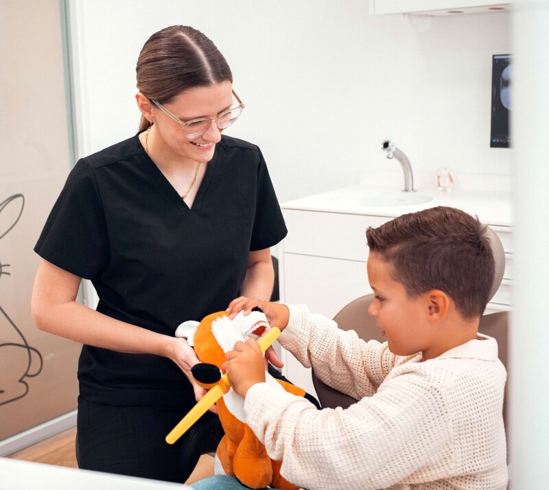 Frau Riekert zeigt einem kleinen Jungen im Behandlungszimmer anhand einer Puppe, wie man die Zähne richtig putzt. Der Junge putzt der Puppe mit einer großen Zahnbürste die Zähne.